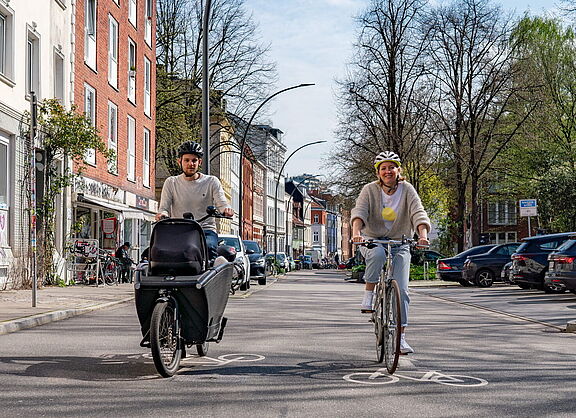 Zwei Personen fahren mit einem Lastenrad mit Transportbox und einem Cityrad auf einer Straße mit Rad-Markierungen; rechts stehen parkende Autos.